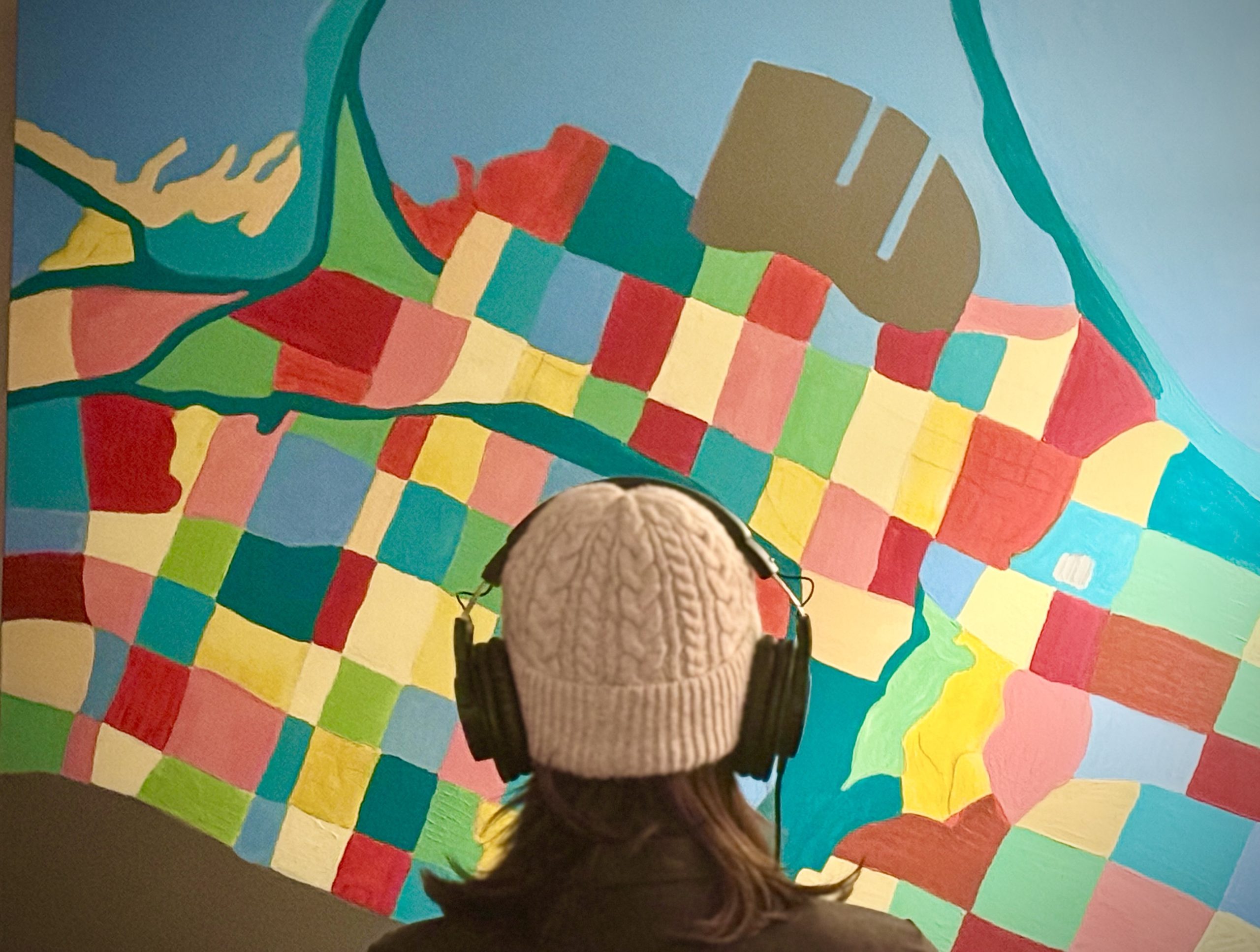 A person wearing headphones is seen from behind, just from the shoulders up. They are facing a colourful painted map of the city of Hamilton, Ontario, Canada.